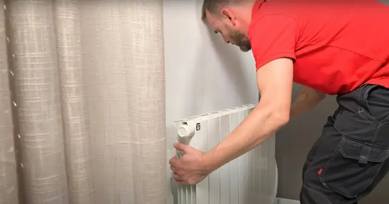Un homme en t-shirt rouge et pantalon de travail ajuste un radiateur électrique blanc, prêt pour son branchement.
